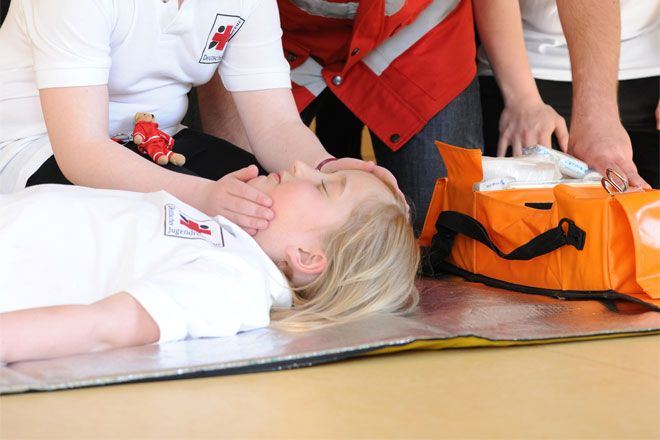 Foto: Bei einer Erste-Hilfe-Übung des Jugendrotkreuzes liegt ein Kind bewusstlos am Boden. Ein anderes Kind überprüft gerade die Atmung. Am Rande des Fotos ist eine Tasche mit Verbandsmaterial zu sehen.