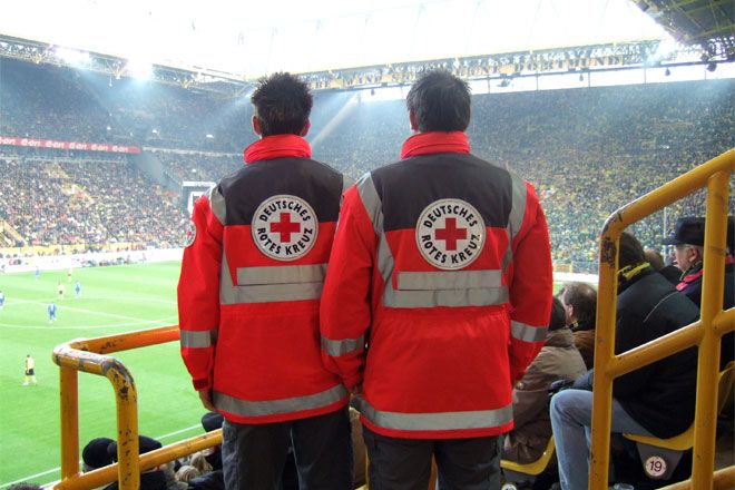 Foto: Mitarbeiter des DRK-Sanitätsdienstes blicken auf das Spielfeld im Stadion.